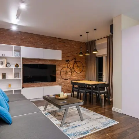 Open-plan living and dining space with an exposed brick feature wall, white TV shelving, and industrial pendant lighting over the dining table.