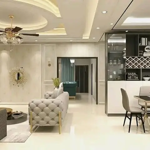 Open-plan living and dining space with an ornate cove-lit stepped false ceiling, tufted gold-leg sofas, and a glass-front bar unit beside the dining table.
