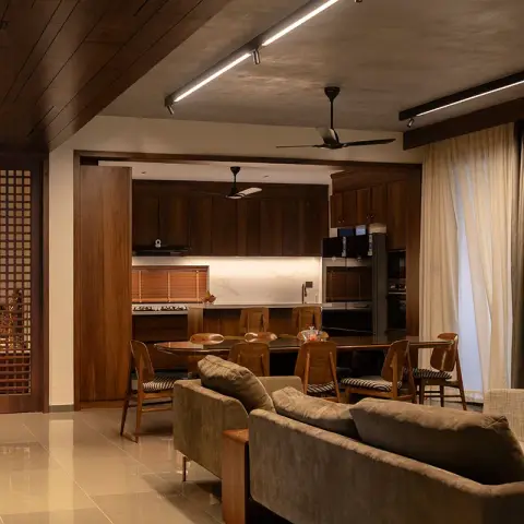 Open-plan living-dining-kitchen with a dark-wood jaali lattice door on the left, a wood-panelled ceiling, dining chairs around an oval table