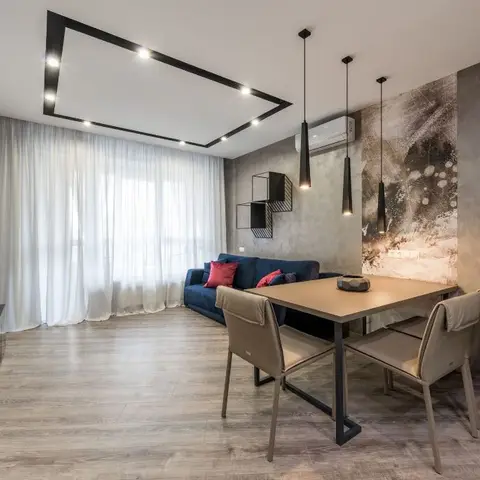 Open-plan living-dining space with a geometric black-trimmed cove ceiling, a textured concrete accent wall, sheer curtains over full-height windows