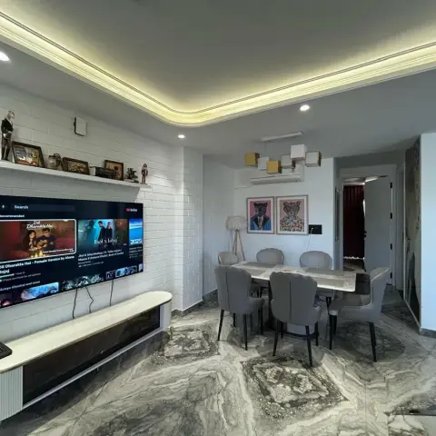 Open-plan living-dining space with a white subway-tile TV wall, cove-lit ceiling, a marble-top dining table with grey upholstered chairs