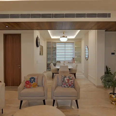 Open-plan living-dining space with floral-embroidered accent chairs in front and a marble dining table beneath a crystal pendant light with glass-front