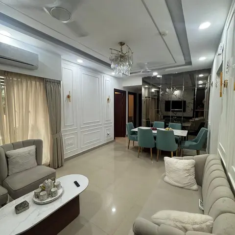 Open-plan living-dining space with a white raised-panel moulding wall, brass sconces, a crystal chandelier over a teal-chair dining table