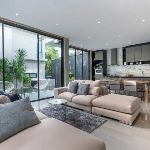 Open-plan living-kitchen-dining space with a beige sectional sofa and grey rug in the foreground, full-height sliding glass doors to a courtyard