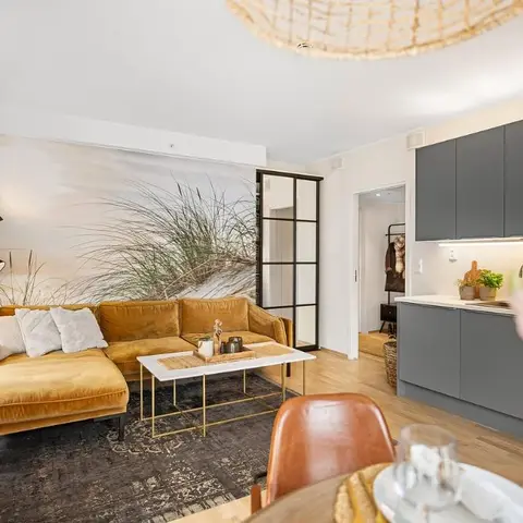 Open-plan living-kitchen-dining space with a mustard velvet sofa against a dune-grass wall mural, a matte grey handleless kitchen