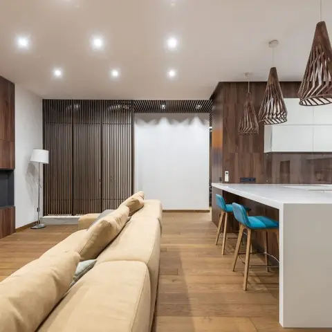 Open-plan living-kitchen with a walnut-panelled TV wall, beige sofa, and three sculptural woven pendant lights over a white waterfall island