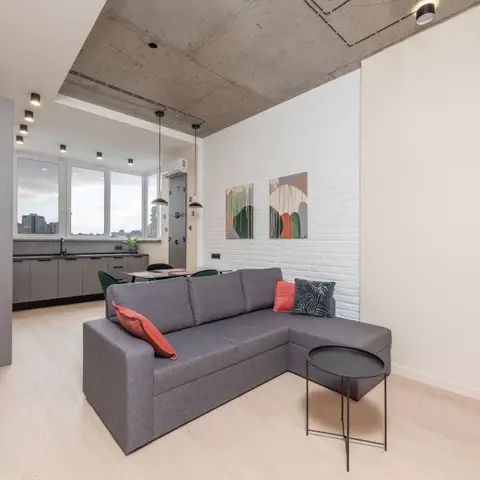 Open-plan living room with a grey L-shaped sofa against a white-painted brick accent wall hung with two framed prints, exposed concrete ceiling beyond.