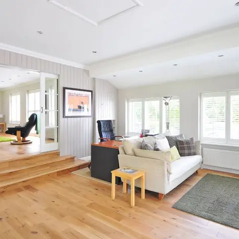 Open-plan living room with a wide-plank pale wood floor, dove-grey wainscot paneling, and a black home-office desk beside glass sliding doors to the next room.
