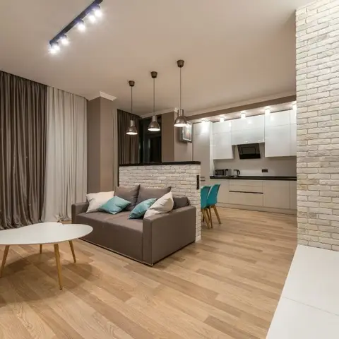 Open-plan living space with a grey sofa and teal accent cushions in front of a white brick-clad partition, kitchen visible beyond with cluster pendant lights.