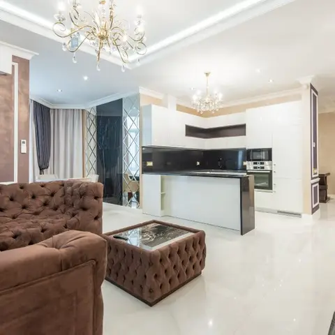Open-plan living space with a tufted brown velvet chesterfield sofa and matching ottoman, crystal chandeliers, a mirrored diamond-pattern accent wall