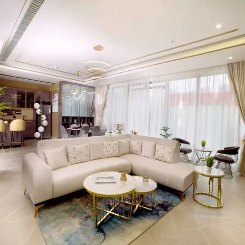 Open-plan living space with a cream sectional sofa and gold nested coffee tables under a ring chandelier, opening onto a dining table and kitchen bar beyond.