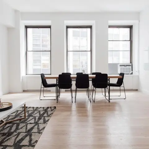 Open-plan loft with a grey sofa and geometric rug in the foreground and an eight-seat black dining table set against a row of three tall windows.