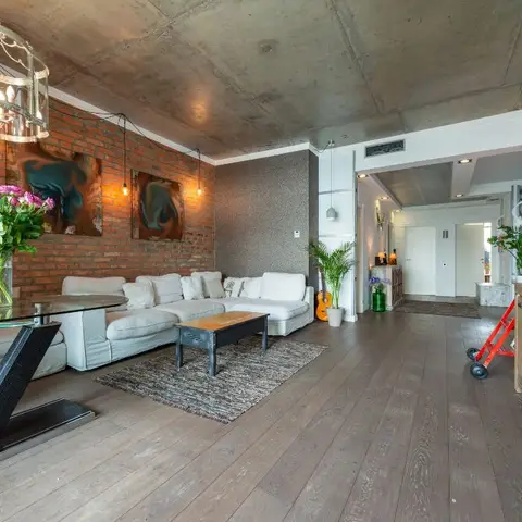 Open-plan loft with an exposed concrete ceiling and brick accent wall, a glass dining table in the foreground and a white sectional sofa opening onto an entry