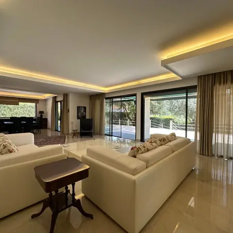 Open-plan lounge and dining area with a cove-lit stepped ceiling, floor-to-ceiling glazing onto a garden terrace, and cream sofas with floral scatter cushions.