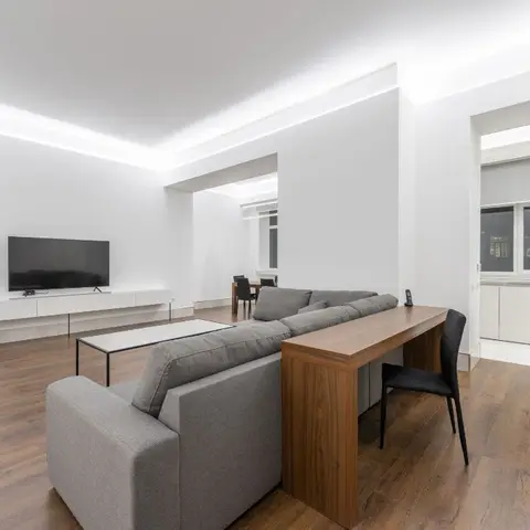 Open-plan minimalist living room with a grey sectional sofa backed by a wood console desk, a floating white TV unit, and a cove-lit ceiling
