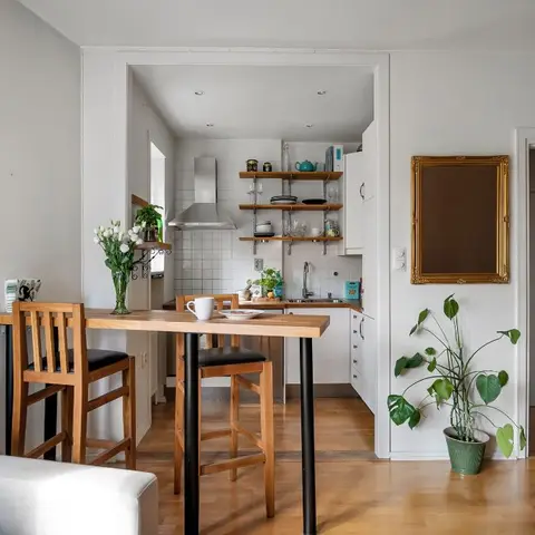 Open-plan scandi apartment with a white-tiled galley kitchen visible through an archway beside a wood high dining table with bar stools