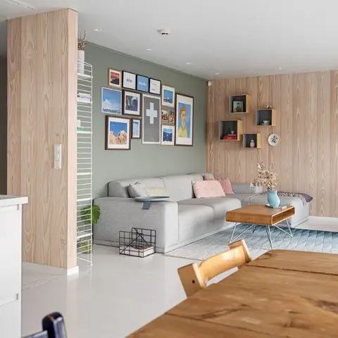 Open-plan Scandinavian space with a wood-panelled accent wall, sage green gallery wall behind a grey sectional, and a wood dining table in the foreground.
