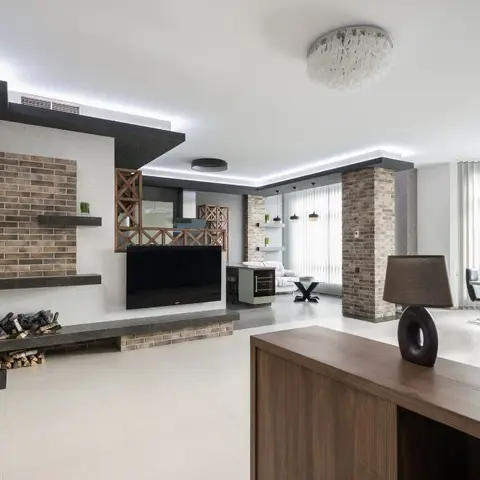 Open-plan space combining a living zone with exposed brick fireplace wall, a kitchen counter, and a dining table, all visible in one wide frame.