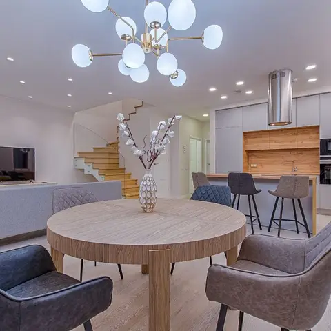 Open-plan space combining a round wood dining table, sofa living zone and galley kitchen with island under one multi-globe chandelier