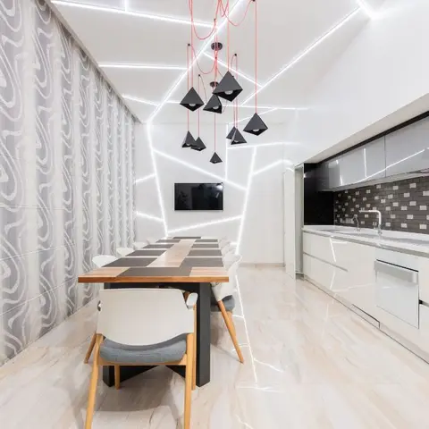 Open-plan space showing a dining table under angular linear-LED ceiling lines with a white galley kitchen and patterned dark backsplash beyond