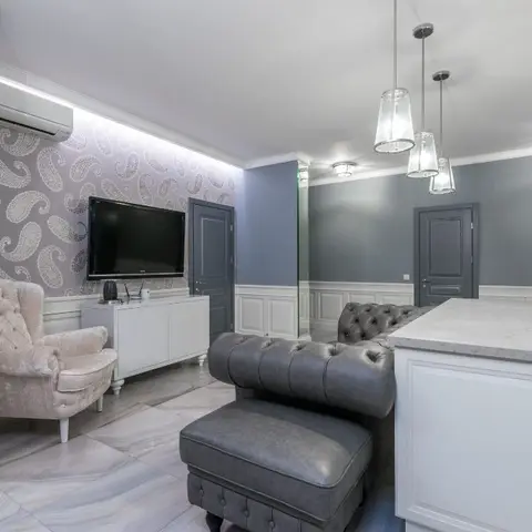Open-plan space with a paisley-wallpapered living zone with a tufted grey chesterfield sofa and armchair, adjoining a white marble kitchen island.