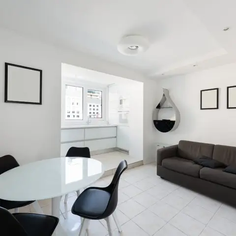Open-plan studio combining a black-and-white dining nook, a brown sofa lounge area, and a glimpse of a small white kitchenette beyond a raised threshold.