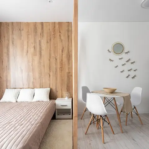 Open-plan studio with a wood-panelled bed nook on one side of a partition wall and a small round dining table with scandi chairs on the other.