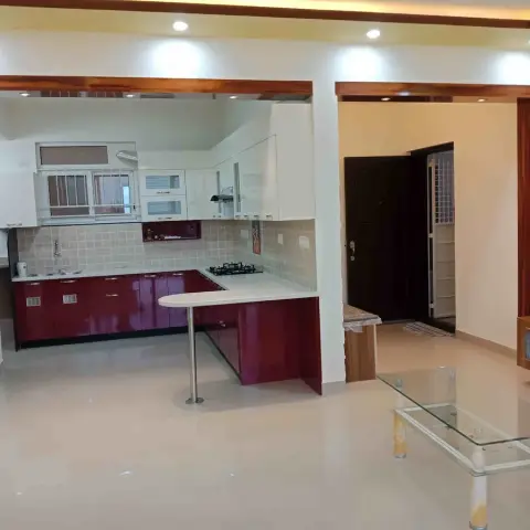 Open-plan view from an L-shaped kitchen with glossy maroon and white cabinetry and a breakfast counter, through to a living area with a wood TV unit