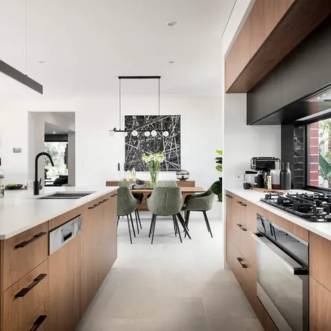 Open-plan walnut-cabinet kitchen island with black gas hob and gooseneck tap, looking through to a dining table with green upholstered chairs under a