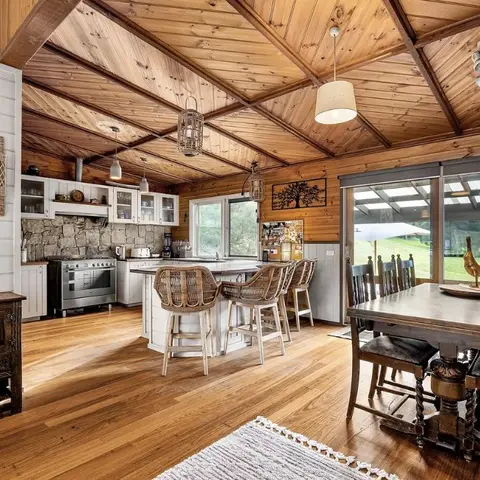 Open-plan wood-ceilinged kitchen with a stone backsplash and wicker bar stools, opening onto a rustic carved dining table beside sliding doors to the garden.