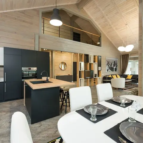 Open-plan wood-clad cabin interior with a dark matte kitchen island, a white dining table set in the foreground and a living seating area with a mezzanine loft