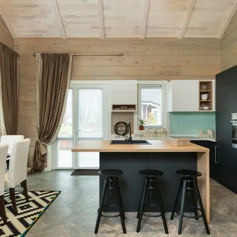 Open-plan wood-clad cabin kitchen with a black island bar seating three, opening directly onto a dining table with slipcovered chairs and a chevron rug.