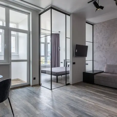 Open studio layout with a grey concrete-textured feature wall behind a wall-mounted TV and sofa, a glass-partitioned bedroom visible beyond