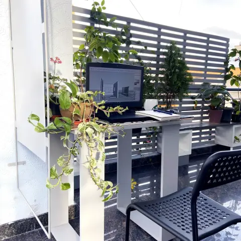 Open terrace turned into a work corner - a white steel desk carrying a laptop with a 3D model on screen, money plant trailing off the shelf beside it