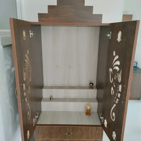 Open wooden pooja unit cabinet with carved brass-inlay door panels and a two-tier idol shelf, installed beside a kitchen counter