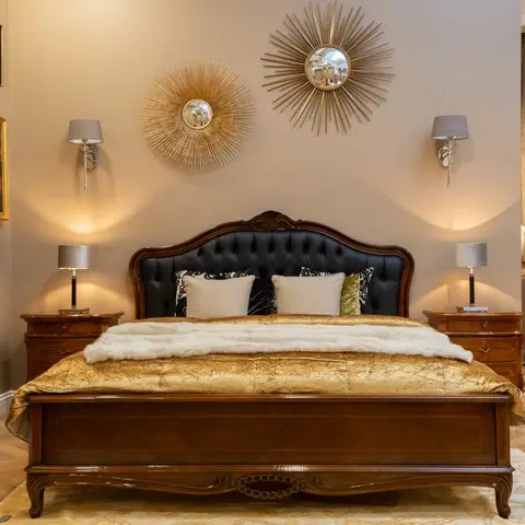 Opulent bedroom with a carved dark wood bed frame and tufted black leather headboard, gold bedding with a fur throw, twin sunburst mirrors