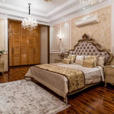 Opulent master bedroom with a gold-trimmed tufted headboard, crystal chandelier, louvered wood wardrobe