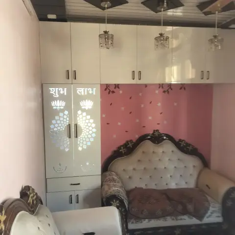 Ornate carved-wood tufted sofa against a pink floral-decal wall, beneath glossy white loft wardrobes with a mirror-work 'Shubh Labh' lotus motif.