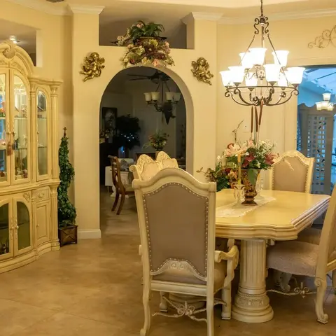 Ornate classic dining room with a carved cream wood table and upholstered nailhead chairs beneath a wrought-iron chandelier