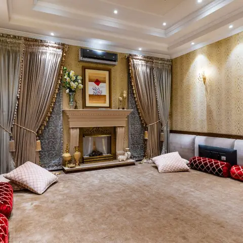 Ornate majlis-style sitting room with gold damask wallpaper, a decorative fireplace mantel, floor-level upholstered seating with red bolster cushions
