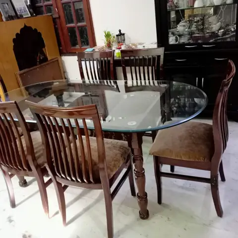 Oval glass-top dining table with six carved wood chairs in mustard velvet cushions, black crockery cabinet behind.