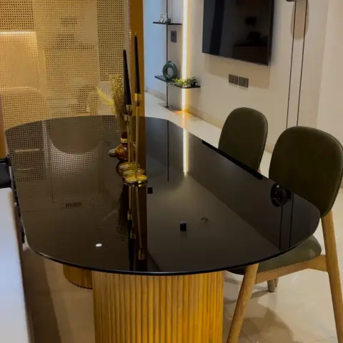 Oval smoked-glass dining top on a fluted teak drum pedestal, set against a backlit perforated brass jaali screen with olive-velvet dining chairs