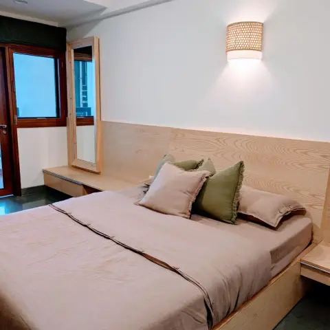 Pale ash platform bed on a wide plinth, with a wood band running the wall behind it into a window-side dressing ledge, olive and dusty-pink linen