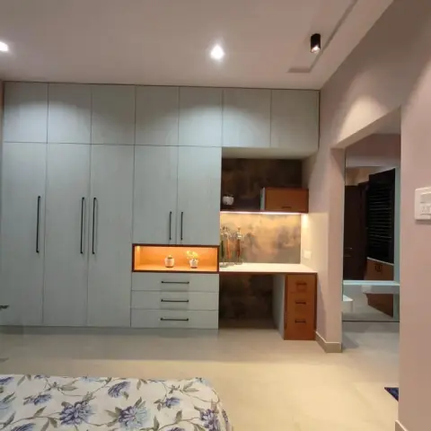 Pale grey-green textured wardrobe wall with a warm-lit open niche and drawer bank, running into a built-in desk set against a copper-patina panel