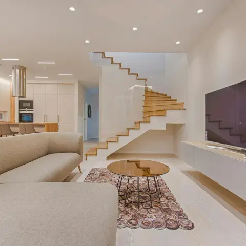 Pale open-plan living zone with an oatmeal L-shaped sofa facing a wall-hung white TV console, an amber glass round coffee table on a circle-pattern rug