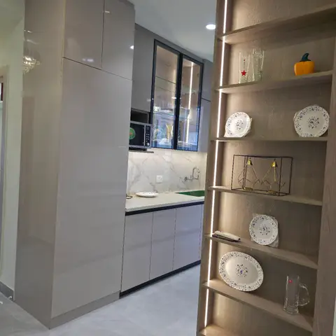 Passage into a grey glossy modular kitchen, framed by a backlit open crockery display shelf styled with plates and glassware.