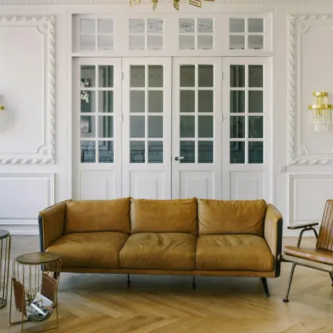 Period apartment living room with a tan leather three-seater centred on herringbone parquet between two brass-and-crystal sconces