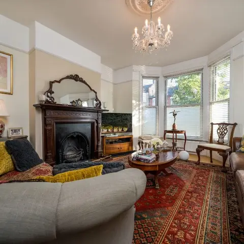 Period living room with a carved wood fireplace mantel, crystal chandelier and a worn Persian rug beneath leather and patterned sofas.