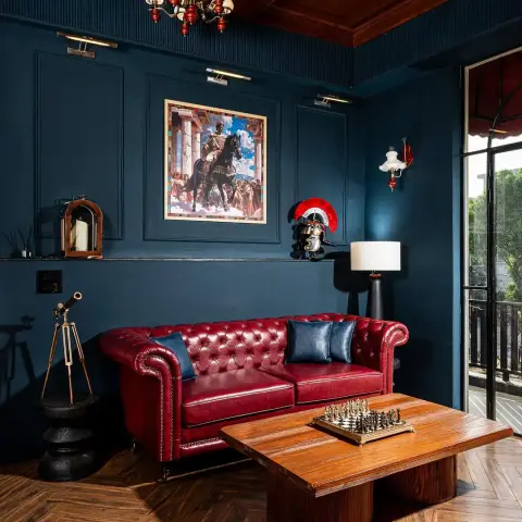 Petrol-blue panelled sitting room with a coffered timber ceiling, an oxblood buttoned Chesterfield under a framed Roman-emperor print