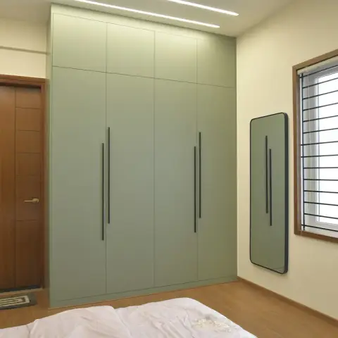 Photo of a bedroom corner with a full-height sage green wardrobe carrying 600mm black tubular handles, a rounded-corner mirror on the adjacent wall